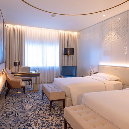 Steigenberger Hotel Ad Dawhah Esterno foto The photo shows a modern hotel room with two single beds placed beside each other. The room features a contemporary design with light-colored walls and stylish decor. There is a large window dressed with sheer curtains, allowing natural light to fill