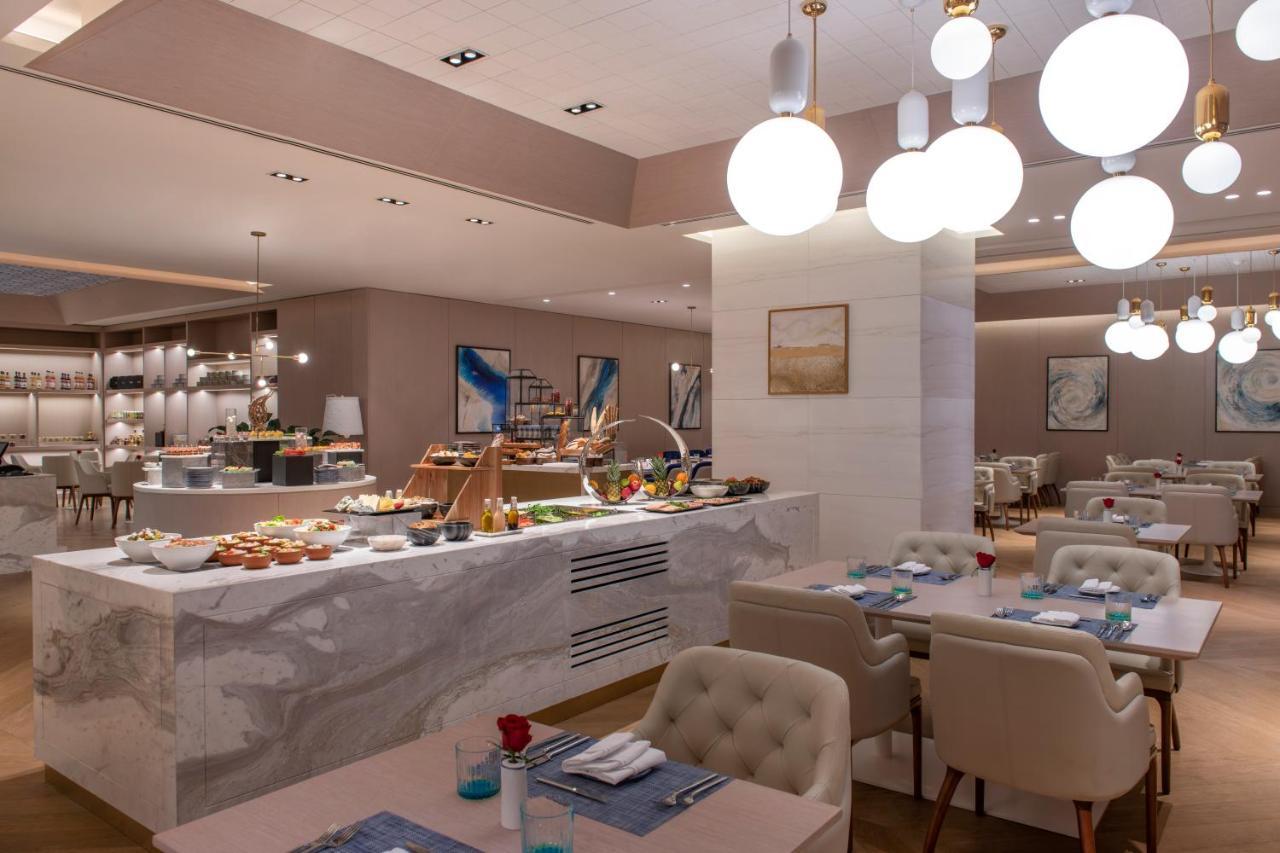 Steigenberger Hotel Ad Dawhah Esterno foto The photo shows a modern restaurant interior that features a stylish and elegant design. In the foreground, there are tables with light-colored upholstered chairs, set with blue napkins and small flower arrangements. 

In the background, there is a b
