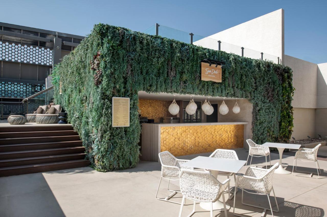 Steigenberger Hotel Ad Dawhah Esterno foto The photo shows an outdoor bar area surrounded by lush greenery. The bar has a modern design, featuring a wooden counter with decorative pendant lights hanging above. There are several white outdoor tables and chairs arranged in front of the bar, whi
