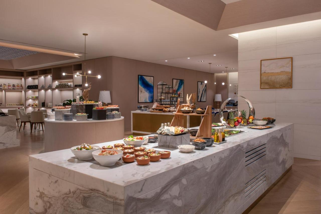 Steigenberger Hotel Ad Dawhah Esterno foto The photo depicts a modern dining area, likely a buffet setup in a restaurant or hotel. A long white marble counter is filled with various dishes in bowls and platters, showcasing an assortment of food items. The space has a sophisticated and elegant