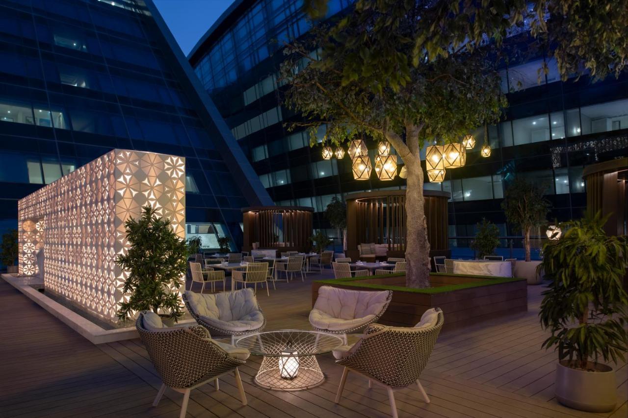 Steigenberger Hotel Ad Dawhah Esterno foto The photo depicts a stylish outdoor terrace area at night. There are modern seating arrangements, featuring woven chairs and a decorative table, surrounded by lush greenery. A large tree is present, adding to the ambiance. 

On one side, there's a st