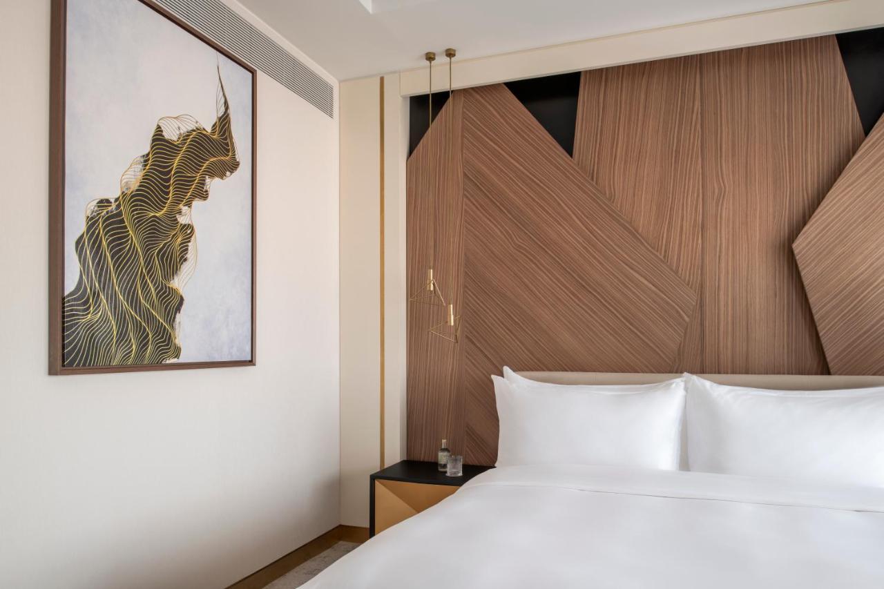 Steigenberger Hotel Ad Dawhah Esterno foto The photo shows a modern hotel room with a clean and stylish design. The focal point is a large bed with white linens, set against a wall that features contemporary wooden paneling with geometric shapes. On the left side, there is a piece of artwork 