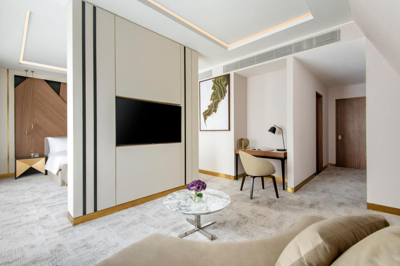 Steigenberger Hotel Ad Dawhah Esterno foto The photo depicts a modern hotel room or suite featuring sleek interior design. It includes a light-colored wall with vertical stripes, a mounted flat-screen television, and a comfortable seating area. A stylish glass coffee table with a small vase o