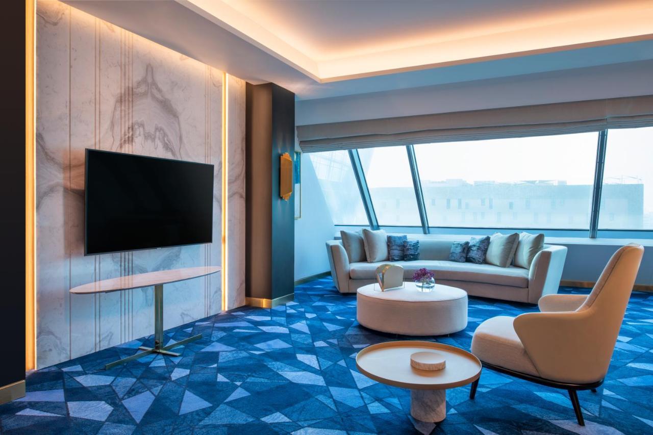 Steigenberger Hotel Ad Dawhah Esterno foto The photo shows a modern and stylish living room with a large window that provides plenty of natural light. There is a flat-screen television mounted on a wall, and a comfortable sofa with cushions is positioned nearby. A round coffee table sits in f