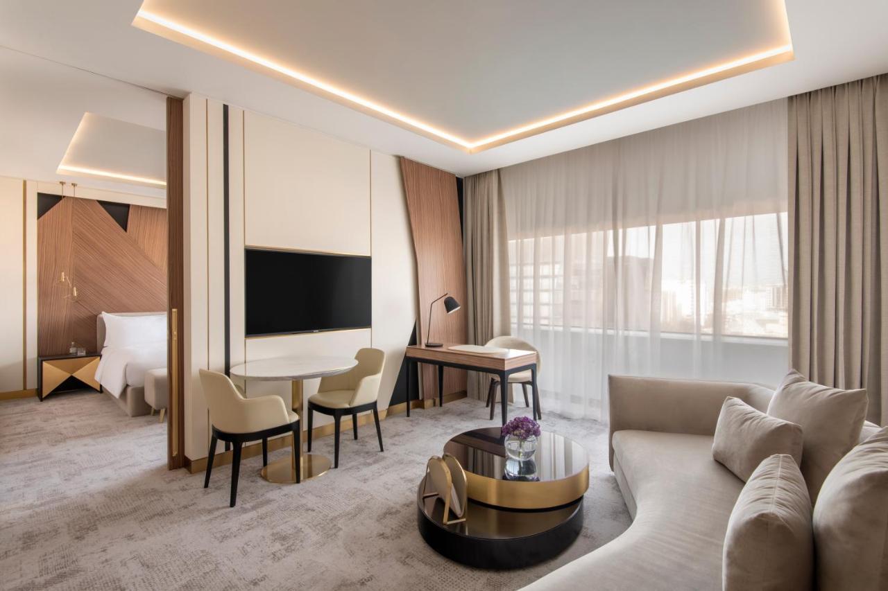 Steigenberger Hotel Ad Dawhah Esterno foto The photo shows a modern hotel room that features a stylish and contemporary design. There is a comfortable seating area with a light-colored sofa and a round coffee table, along with two chairs and a small desk. A large flat-screen television is mou