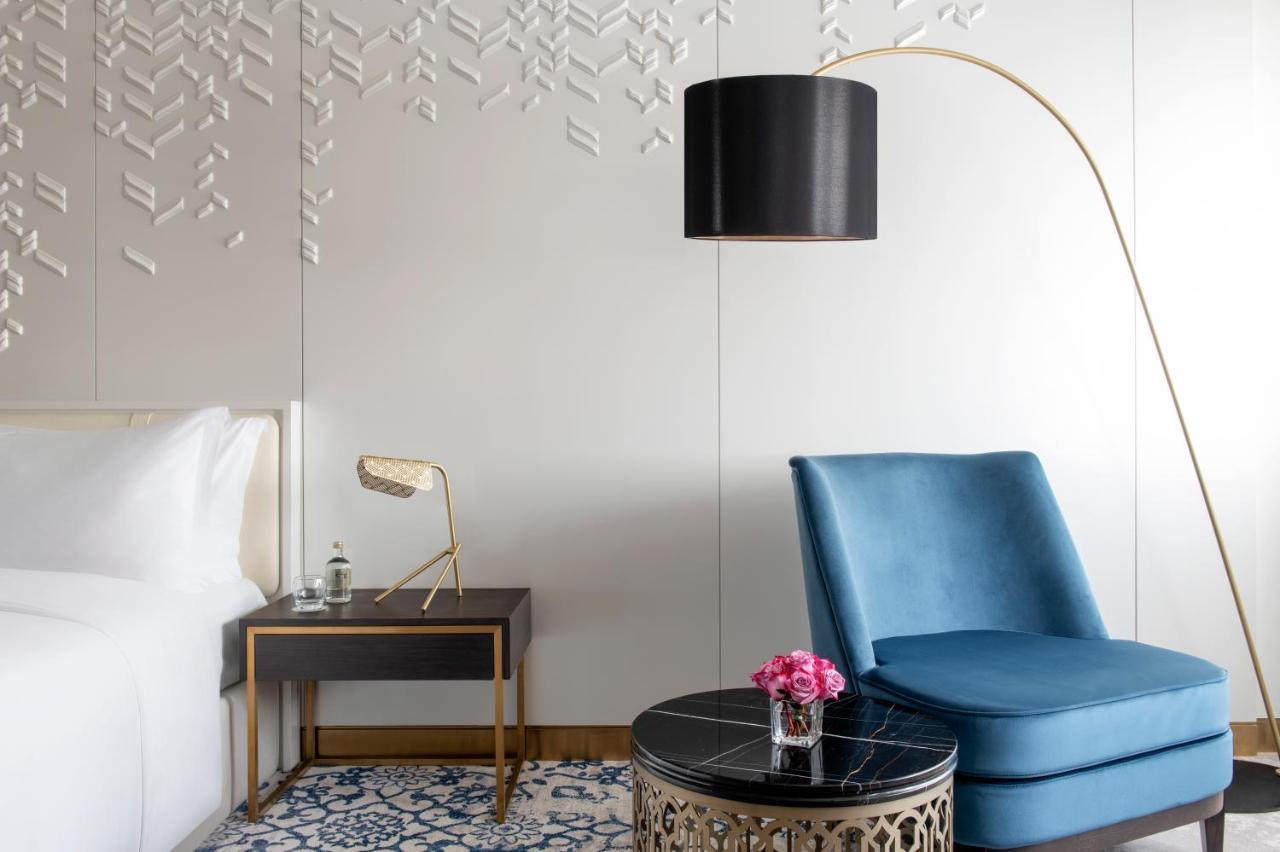 Steigenberger Hotel Ad Dawhah Esterno foto The photo shows a modern interior of a hotel room or living space. On one side, there is a neatly made bed with white linens. Next to the bed, there is a black bedside table with a stylish lamp. 

In the opposite corner, there is a blue accent chair 
