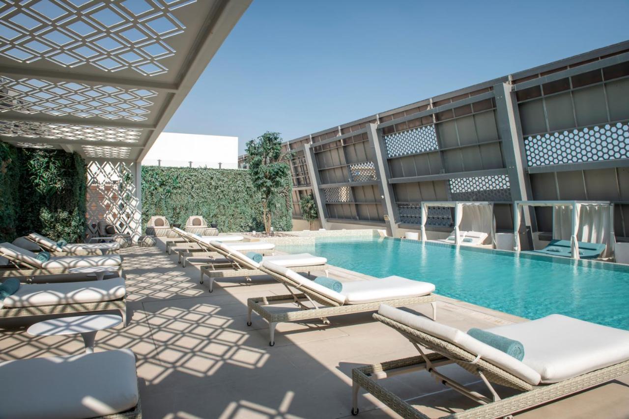 Steigenberger Hotel Ad Dawhah Esterno foto The photo shows a modern outdoor pool area. There are several lounge chairs arranged around the pool, which is a vibrant blue color. Sunlight casts intricate shadows on the ground due to a lattice structure overhead. On one side, there are green plan