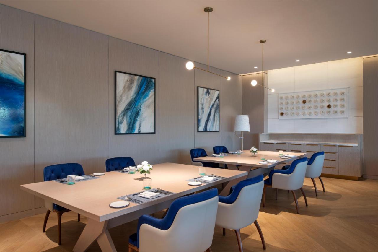Steigenberger Hotel Ad Dawhah Esterno foto The photo shows a modern, elegant dining or meeting room. It features a large, light-colored rectangular table surrounded by chairs with blue upholstery and white frames. Above the table, there are two hanging pendant lights providing illumination. T