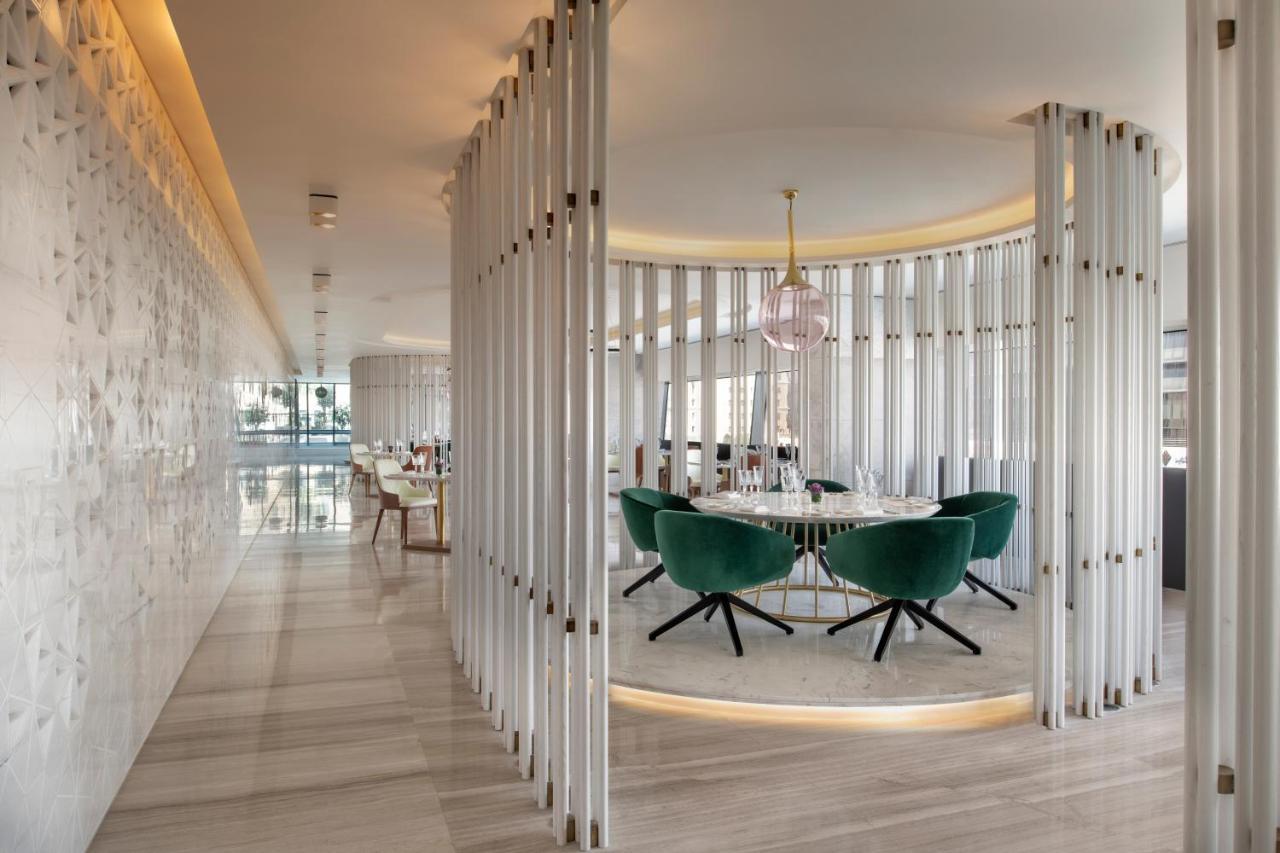 Steigenberger Hotel Ad Dawhah Esterno foto The photo shows a modern, stylish interior of a restaurant or dining area. There is a circular dining table surrounded by green upholstered chairs with a sleek design. The space is framed by vertical white panels, creating a sense of separation and p