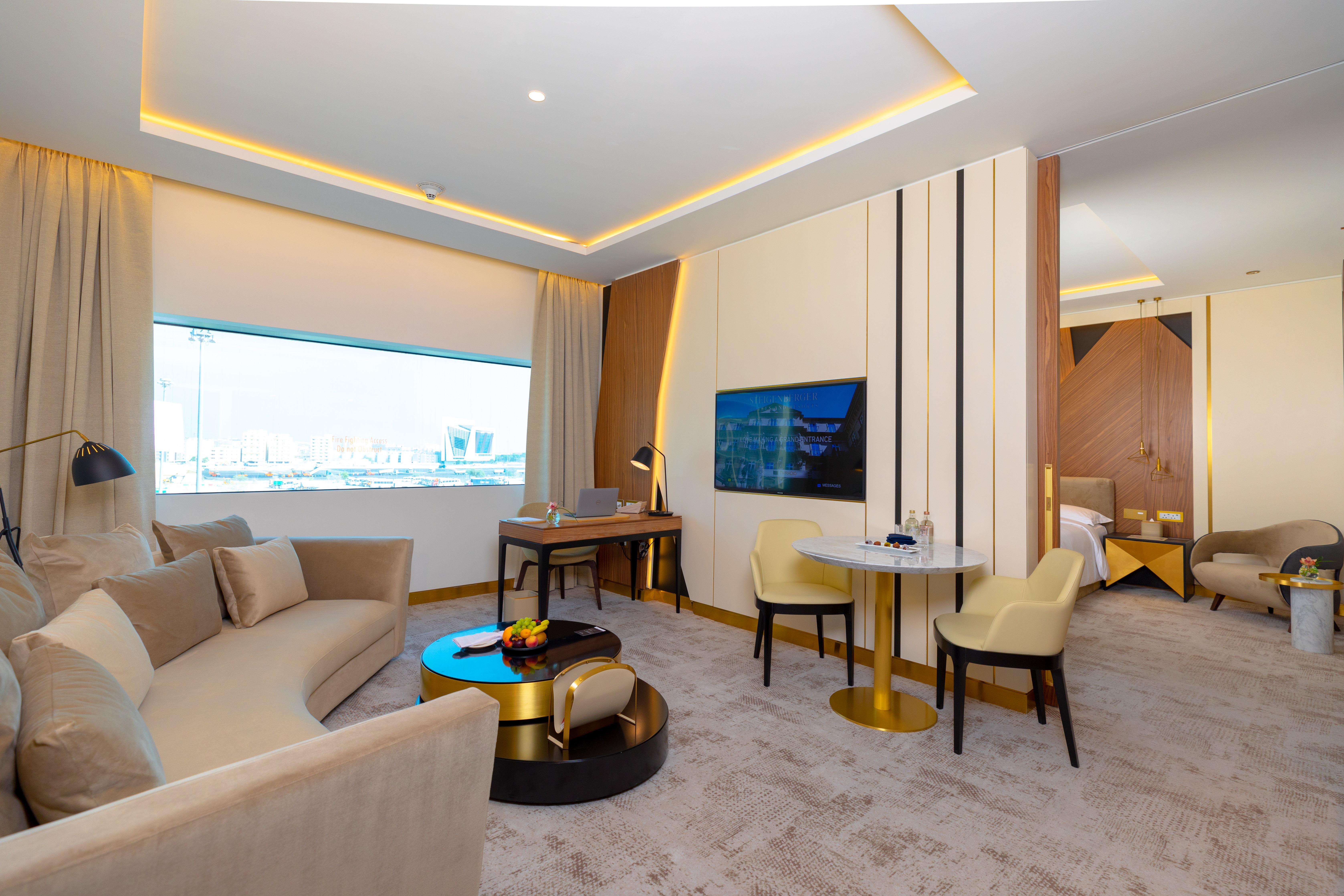 Steigenberger Hotel Ad Dawhah Esterno foto The photo shows a modern and stylish hotel suite. It features a spacious living area with a curved sofa set and a small round table accompanied by two chairs. There is a large window that allows natural light to fill the room, enhancing its bright an