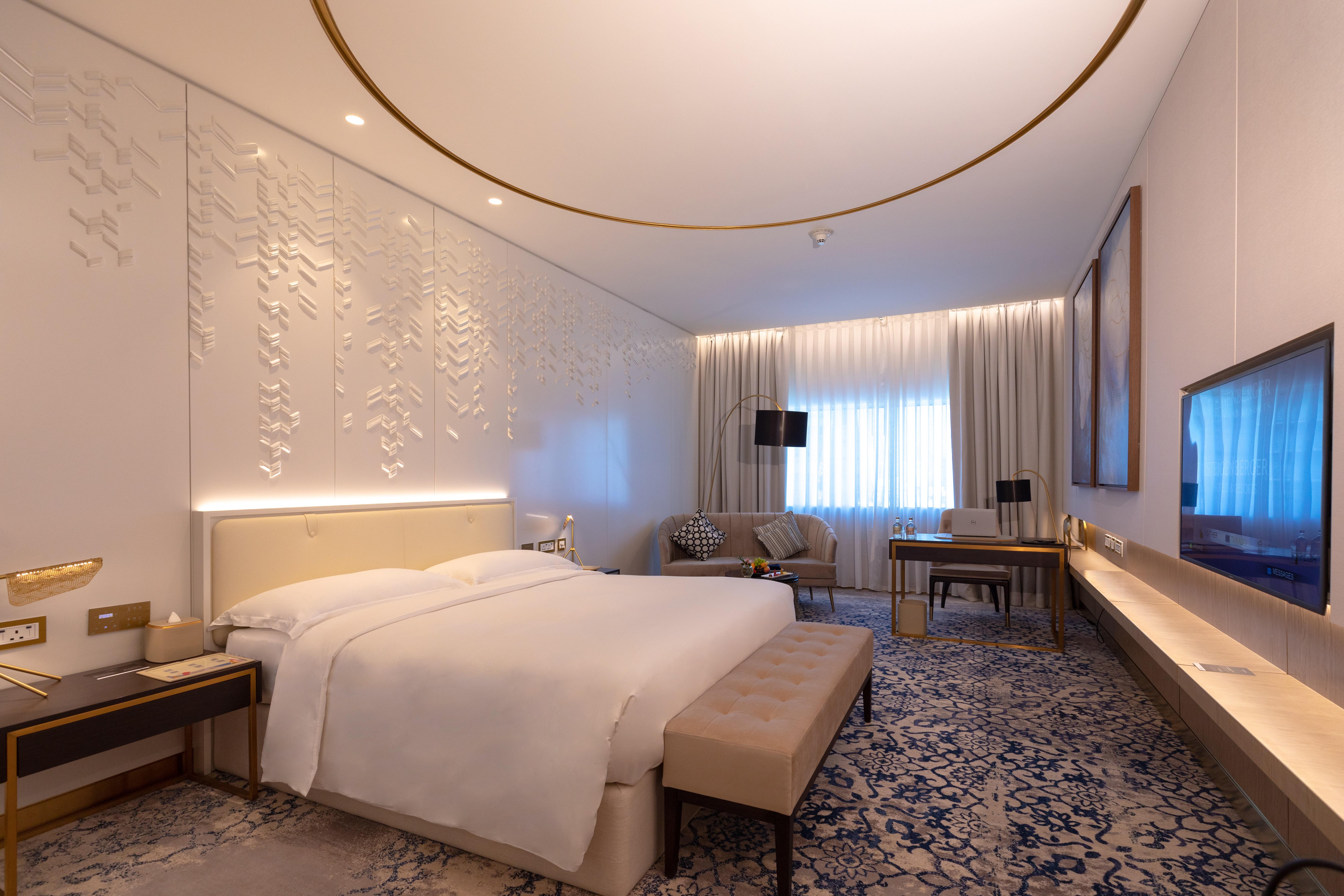 Steigenberger Hotel Ad Dawhah Esterno foto The photo shows a modern hotel room designed with a contemporary aesthetic. It features a large bed with white linens, a soft beige bench at the foot of the bed, and elegant furnishings including a bedside table and a comfortable seating area with ch