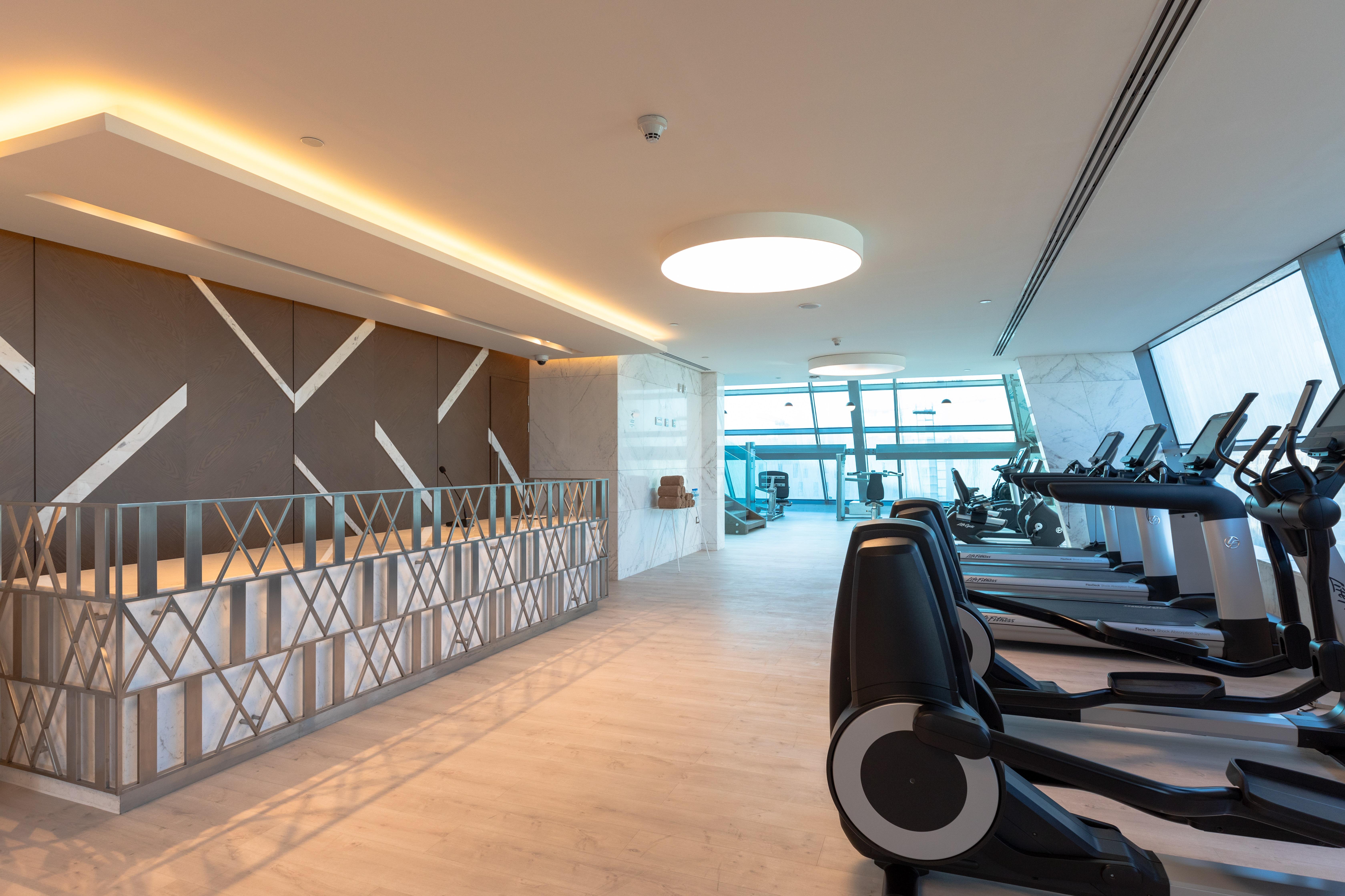 Steigenberger Hotel Ad Dawhah Esterno foto The photo shows the interior of a modern gym. On the left side, there is a reception area with a stylish design featuring geometric patterns on the wall. The area is well-lit with natural light coming in from large windows, providing a view outside. 