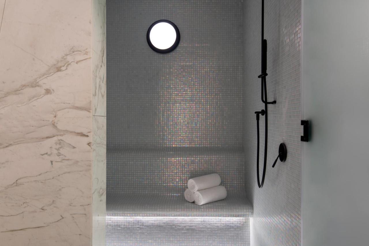 Steigenberger Hotel Ad Dawhah Esterno foto The photo features a modern shower space. The walls are adorned with small mosaic tiles that create a sleek and reflective surface. There is a round window that allows natural light to enter, enhancing the ambiance. A black shower head and faucet are