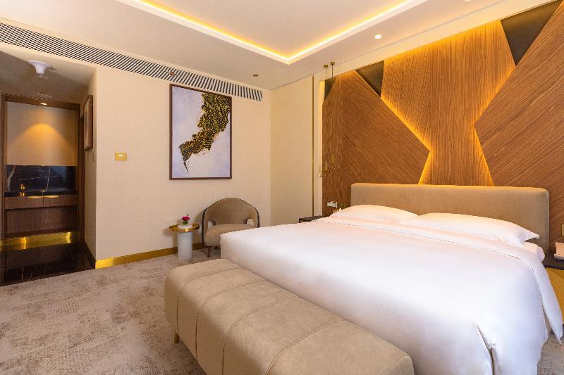 Steigenberger Hotel Ad Dawhah Esterno foto The photo shows a modern hotel room featuring a comfortable bed with a plush white duvet, flanked by elegant decorative elements. The headboard has a stylish wood grain design, accompanied by warm lighting that creates a cozy atmosphere. A small seat